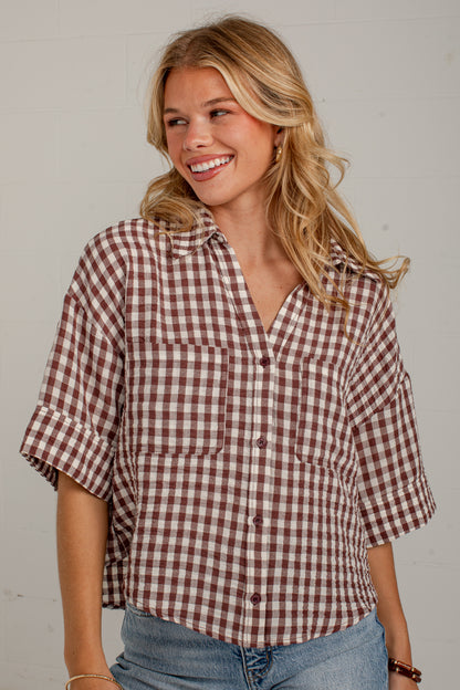 A smiling blonde woman wearing a brown and white gingham check short-sleeve button-up shirt with rolled cuffs and blue jeans, standing against a light gray wall.