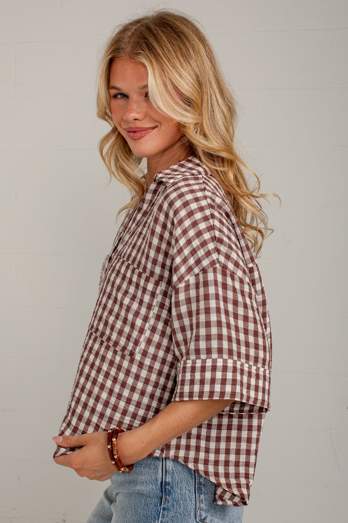 A woman wearing a brown and white gingham check shirt with rolled-up sleeves, paired with light blue jeans, standing against a white paneled wall.
