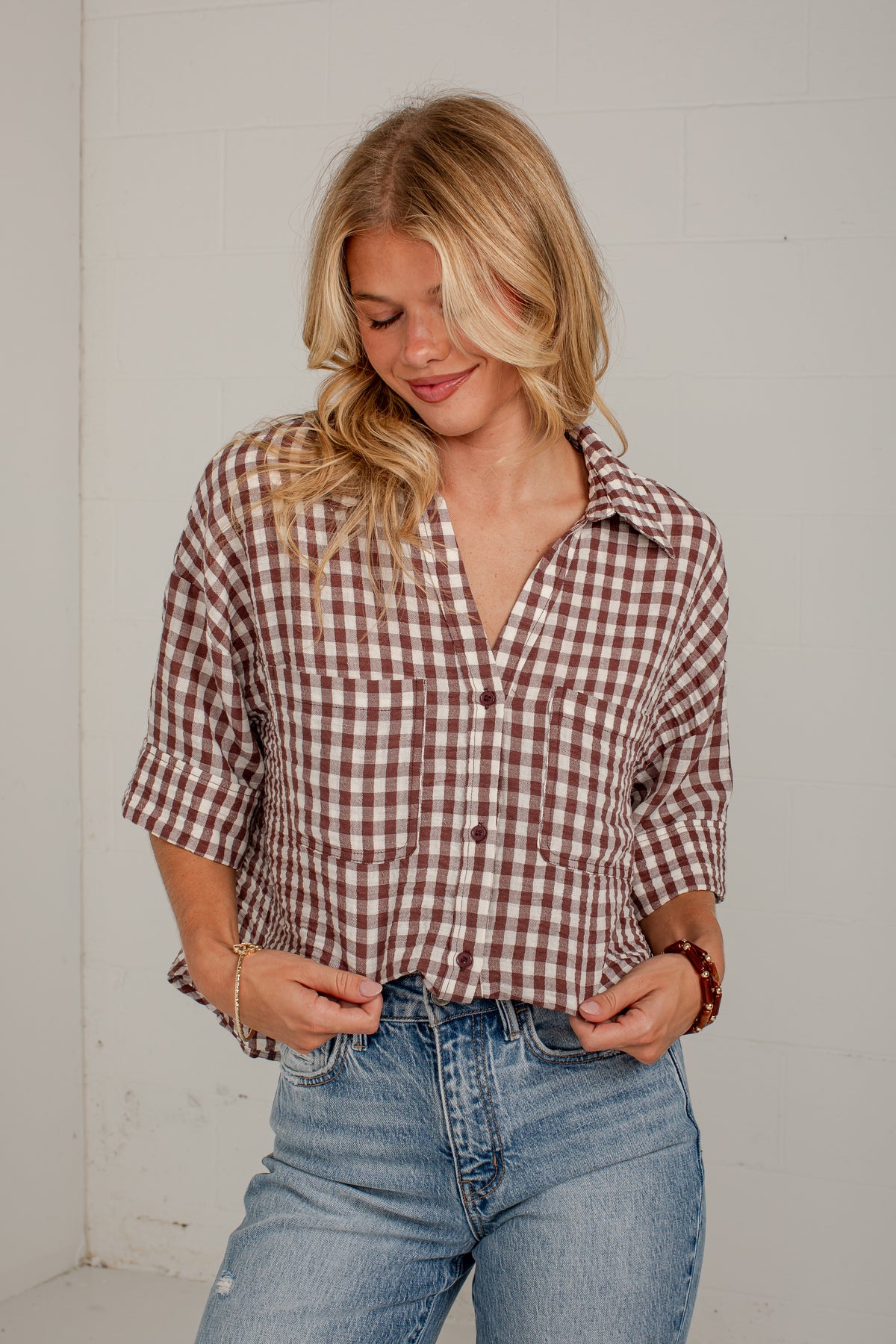 A woman wearing a brown and white gingham plaid button-down shirt with three-quarter sleeves and blue jeans, standing against a white wall.