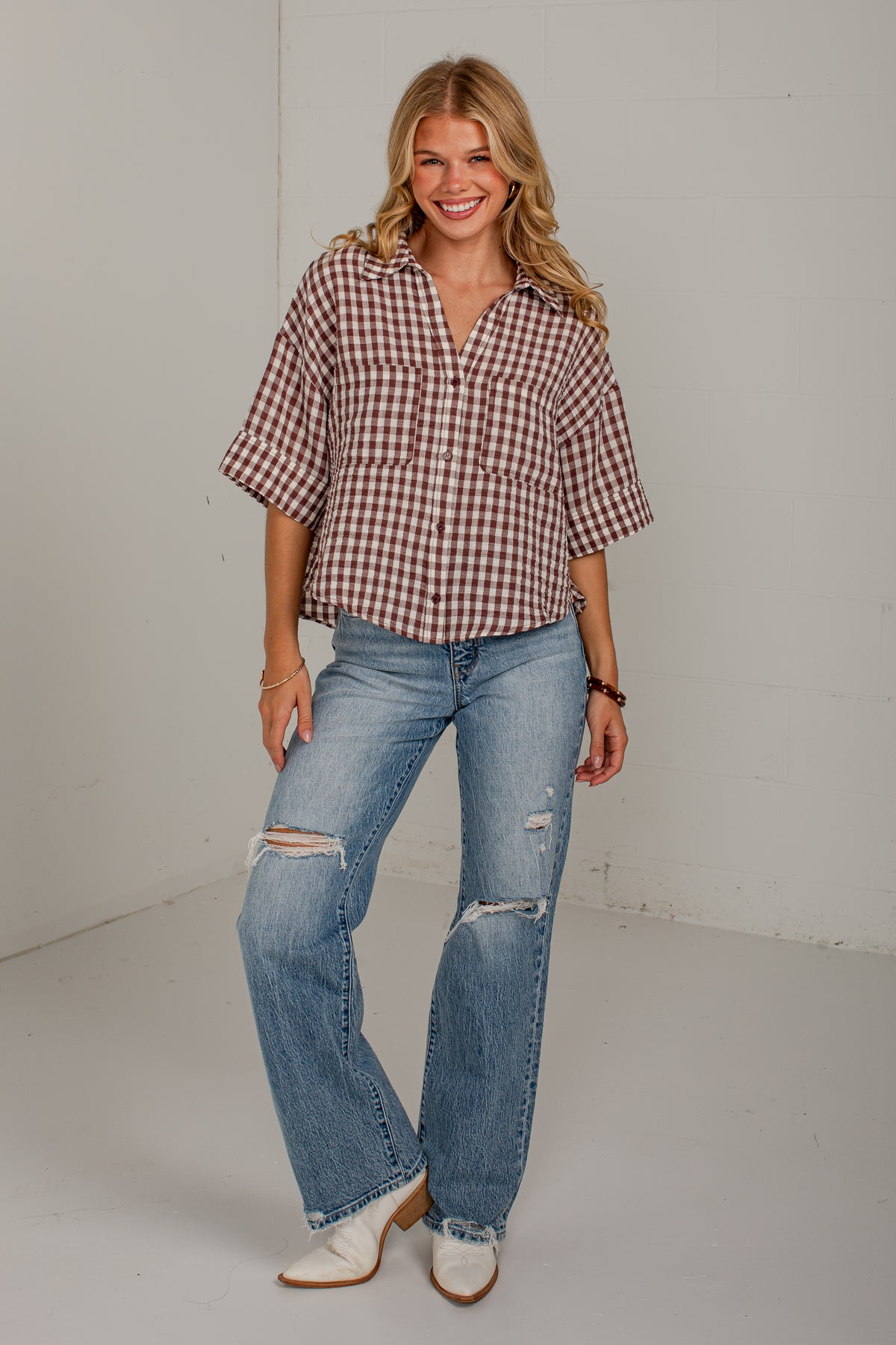 A woman modeling a brown and white gingham short-sleeve button-down shirt paired with light-wash distressed jeans and white cowboy boots, standing against a plain off-white studio wall.