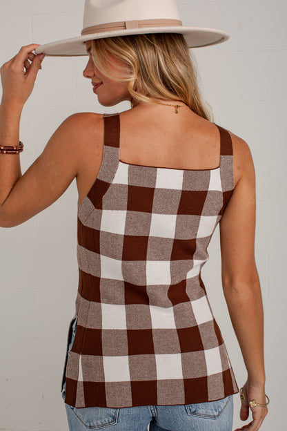 A woman wearing a brown and white buffalo check plaid tank top, blue jeans, and a beige wide-brim hat, shown from a rear three-quarter angle against a white brick wall.