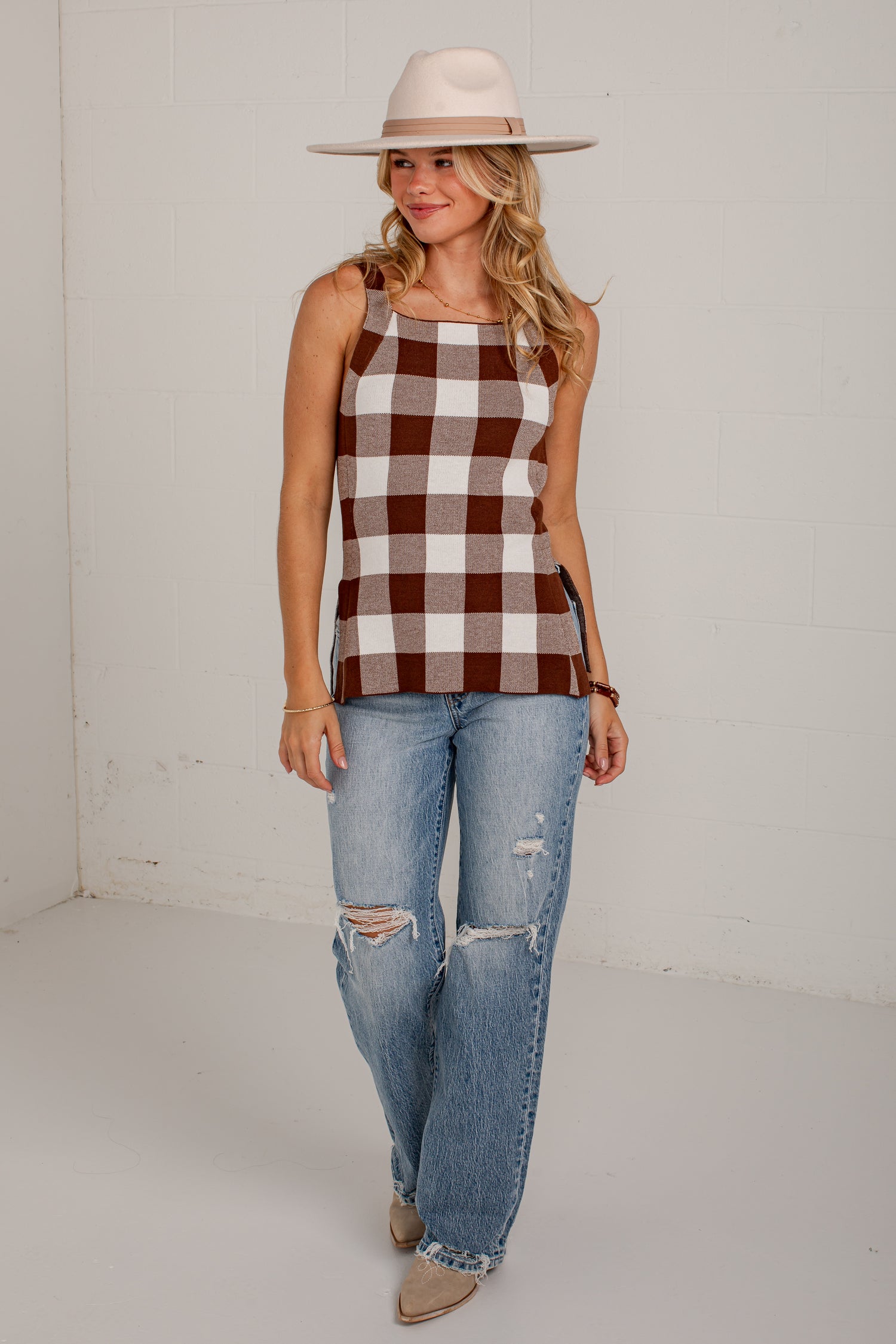 A woman modeling a brown and white buffalo check plaid sleeveless tank top, paired with distressed light-wash blue jeans and a beige wide-brim hat, standing against a white brick wall.