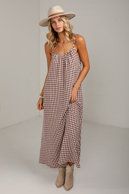A woman modeling a brown and white gingham maxi dress with spaghetti straps, styled with a beige wide-brim hat and beige ankle boots in a bright studio setting.
