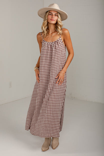 A woman modeling a sleeveless brown and beige gingham maxi dress with a cream wide-brim hat and beige ankle boots, standing in a bright white studio.