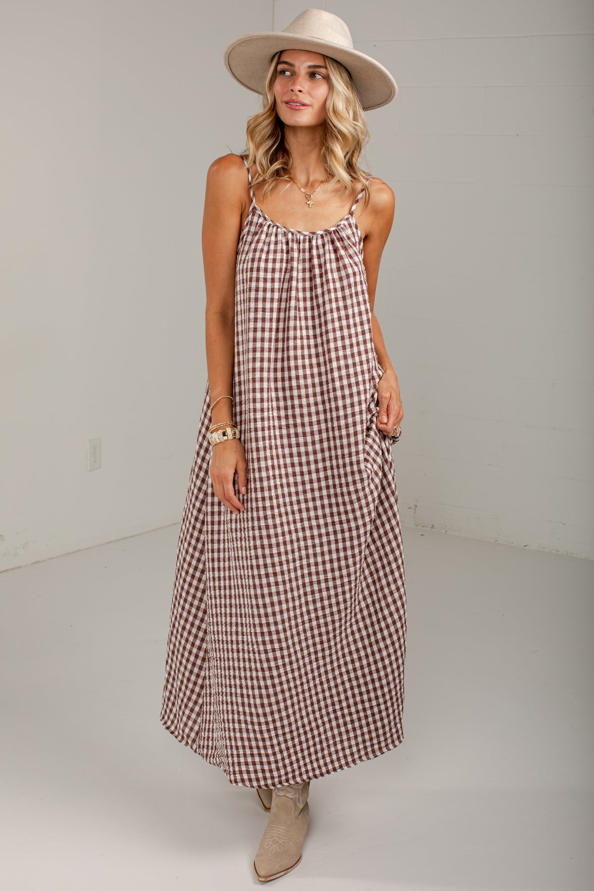 A woman modeling a sleeveless, midi-length gingham dress in brown and white. She is wearing a wide-brimmed beige hat, a gold chain necklace, and beige ankle boots, standing against a plain light gray studio background.