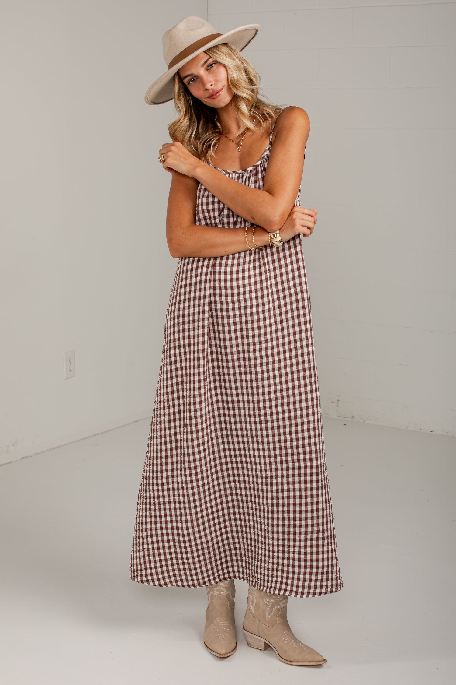 A woman modeling a brown and white gingham maxi dress with spaghetti straps, styled with a beige wide-brim hat, layered gold necklaces, and beige ankle boots against a white studio background.