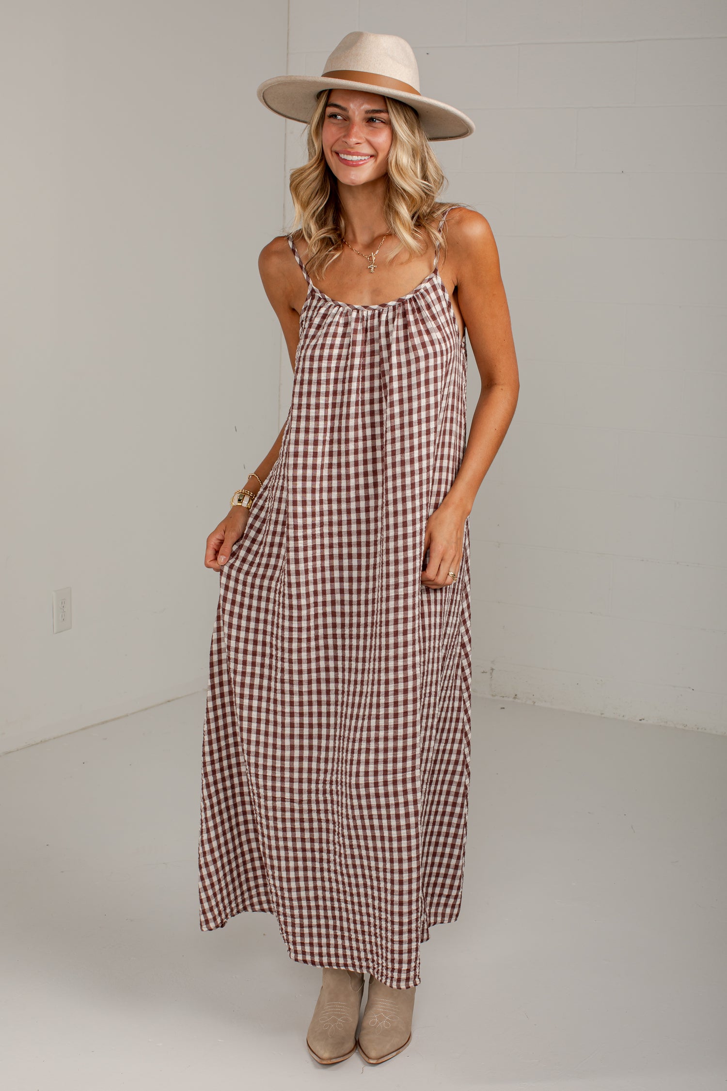 A woman modeling a brown and white gingham maxi dress with spaghetti straps and side pockets, paired with a beige wide-brim hat and beige ankle boots, in a bright white studio setting.