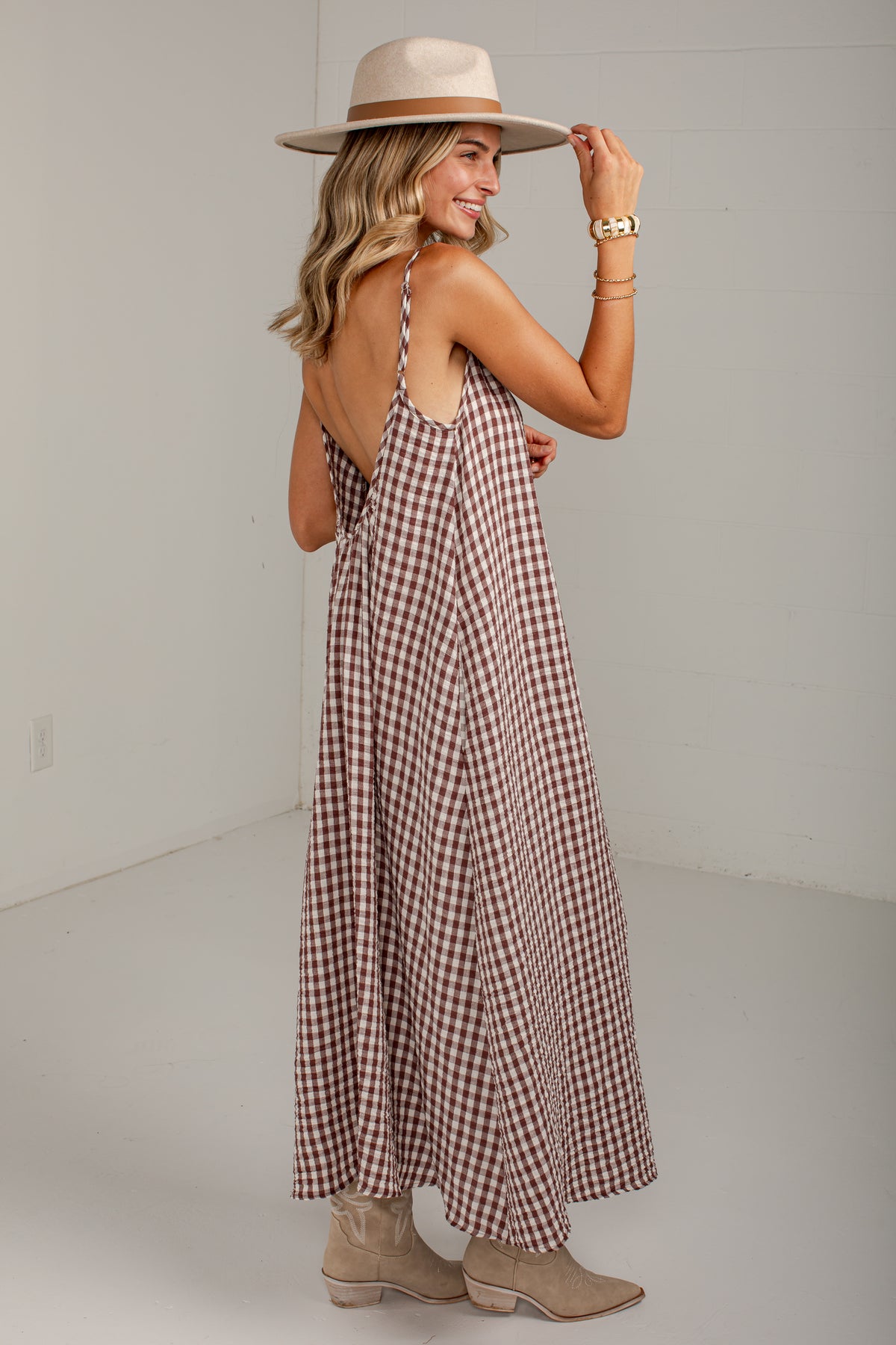 A woman wearing a brown and white gingham maxi dress with a backless open design, styled with a beige wide-brim hat and beige ankle boots, posing in a minimalist studio setting.