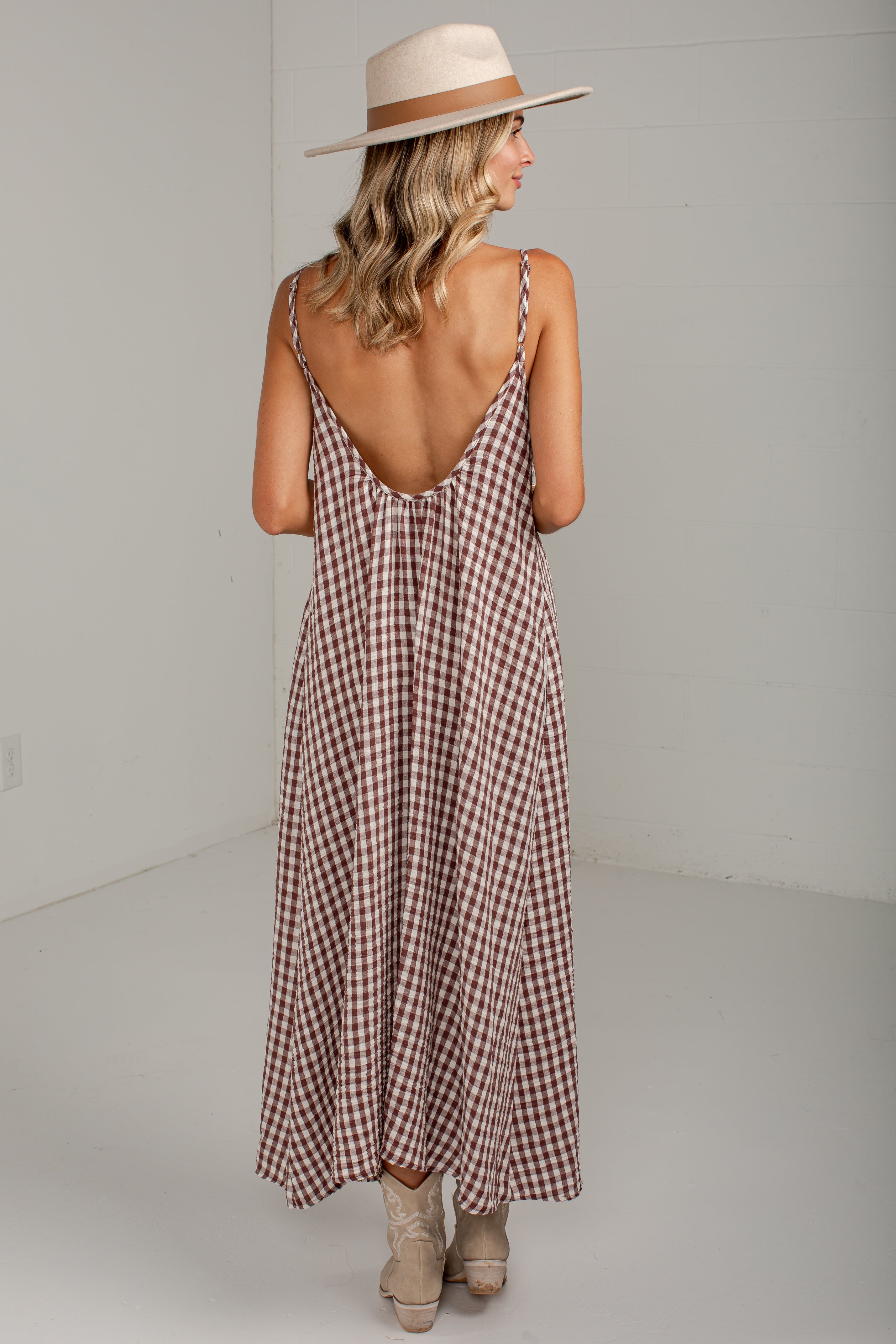 A woman wearing a brown and white gingham maxi dress with a deep open back, styled with a beige wide-brim hat and beige boots, photographed from behind in a minimalist studio setting.