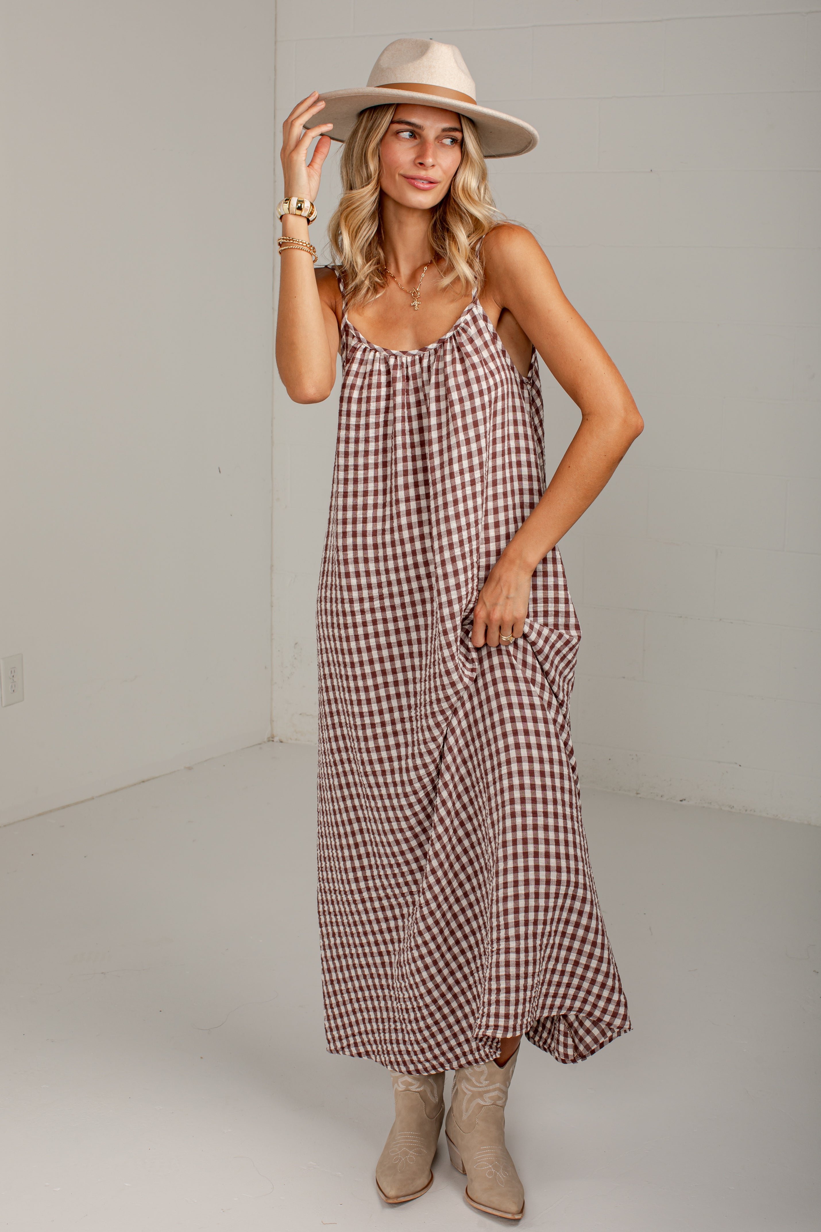 A woman modeling a brown and beige gingham maxi dress with spaghetti straps, a wide-brim beige hat, and beige cowboy boots against a white studio background.