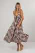 A woman modeling a brown and white gingham midi dress with spaghetti straps and a tie detail at the bust, standing against a light gray studio background.