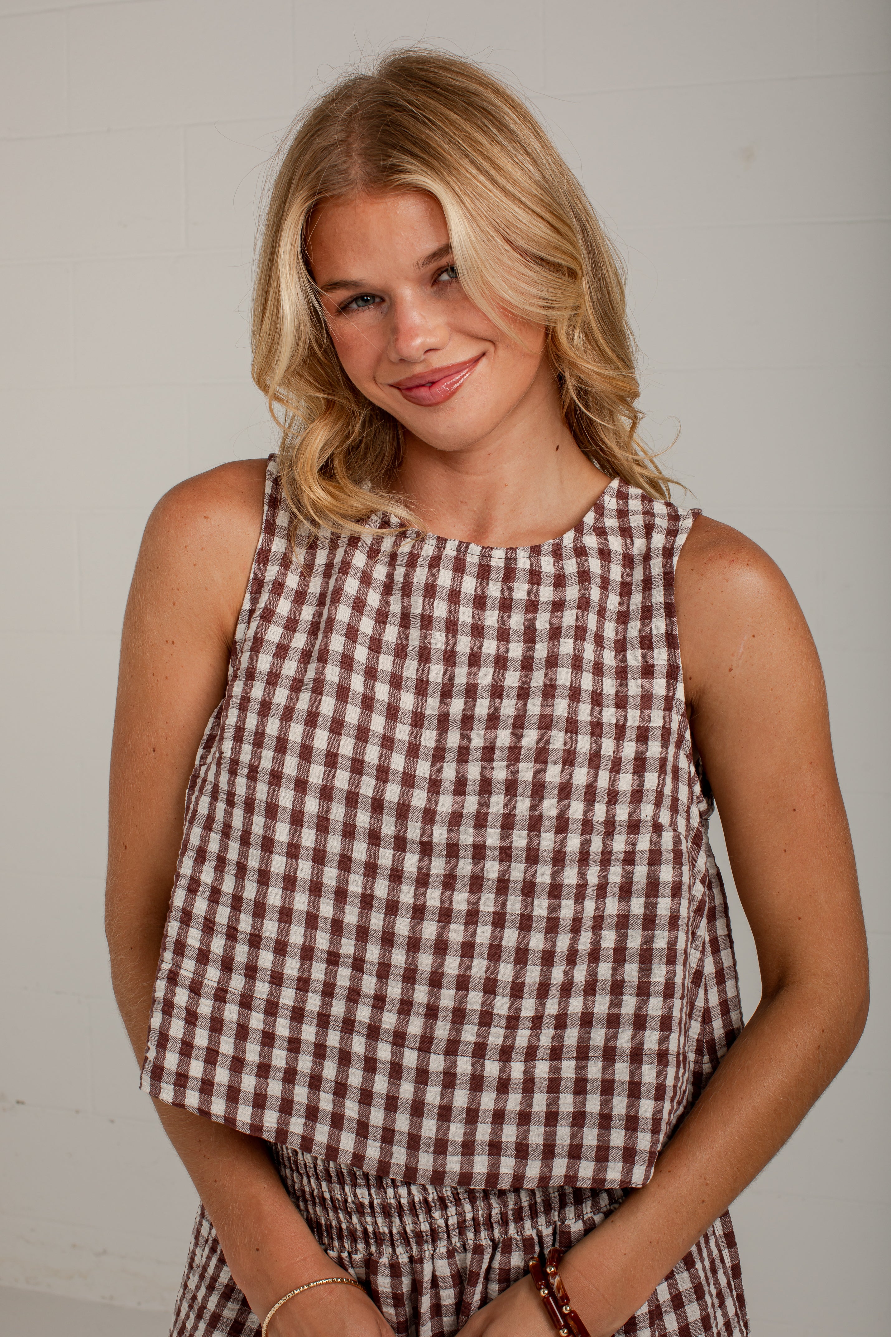 A woman wearing a sleeveless gingham top in brown and white, paired with matching brown gingham bottoms, photographed against a plain light gray wall.