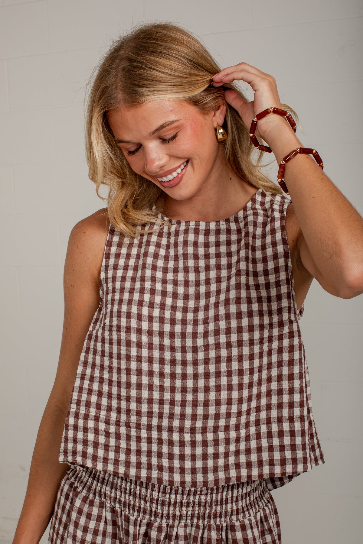 A smiling blonde woman wearing a brown and white gingham check sleeveless top and matching bottoms, accessorized with red beaded bracelets, against a light gray wall.