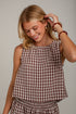 A smiling blonde woman wearing a brown and white gingham check sleeveless top and matching bottoms, accessorized with red beaded bracelets, against a light gray wall.