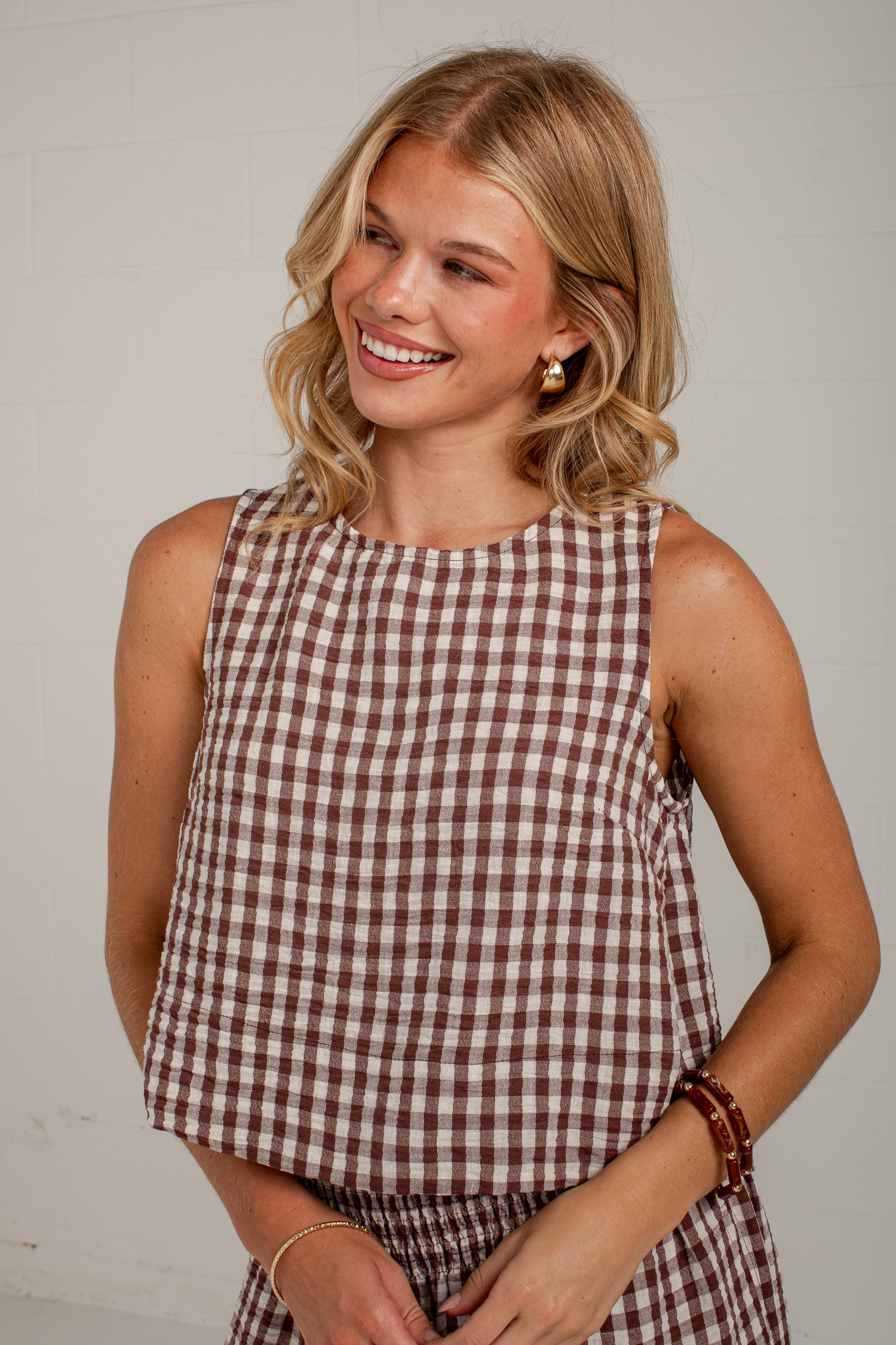 A smiling blonde woman wearing a sleeveless brown and white gingham top, photographed against a plain light gray wall.
