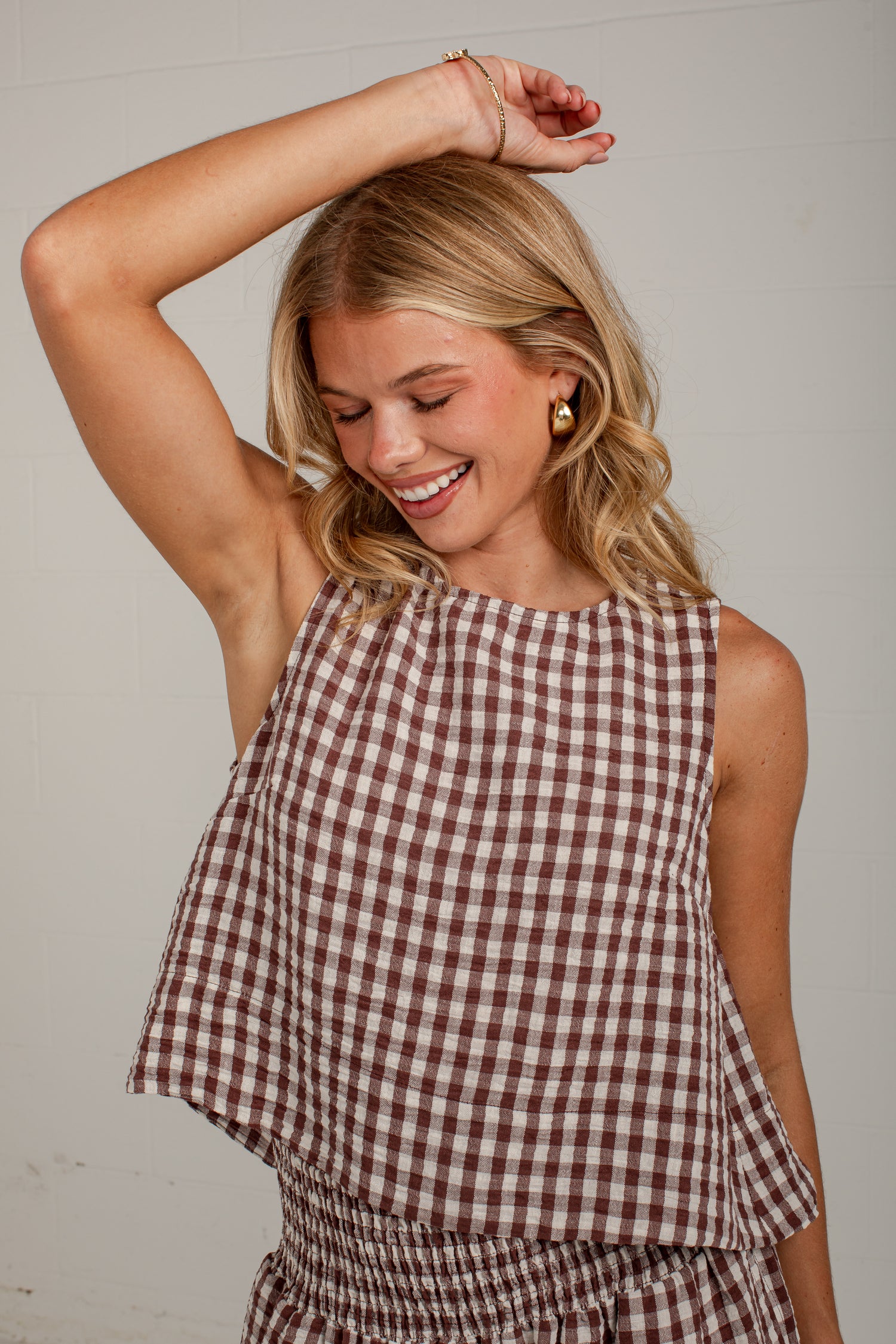 A woman wearing a brown and white gingham sleeveless top and matching bottoms, smiling with one arm raised against a plain light background.