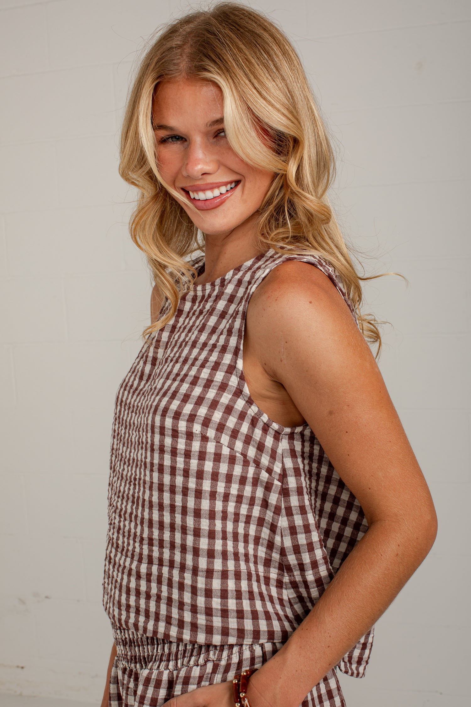 A smiling blonde woman wearing a sleeveless brown and white gingham check romper, posing against a light off-white brick wall.