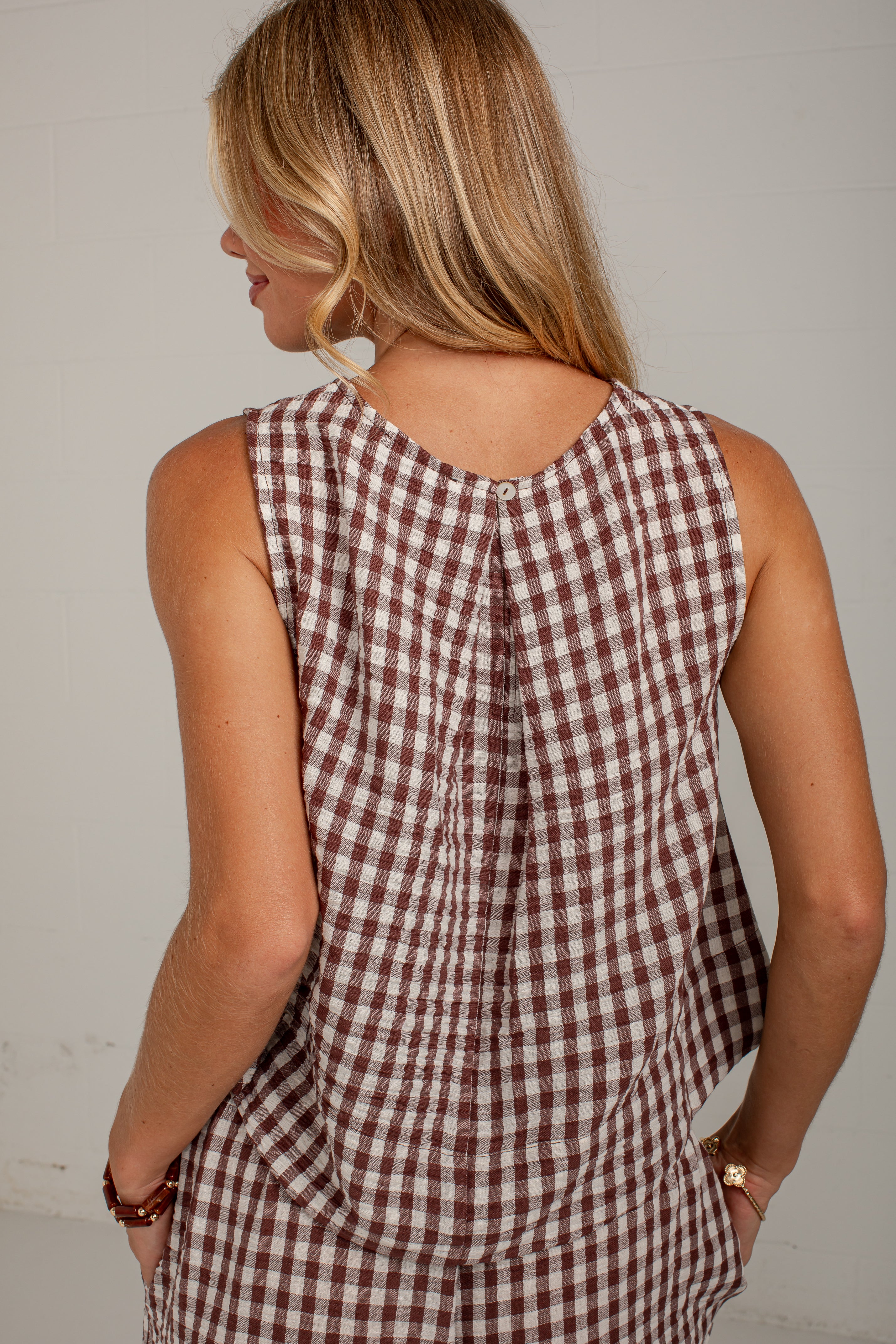 A woman shown from behind wearing a sleeveless brown and white gingham check top, photographed against a plain light wall.