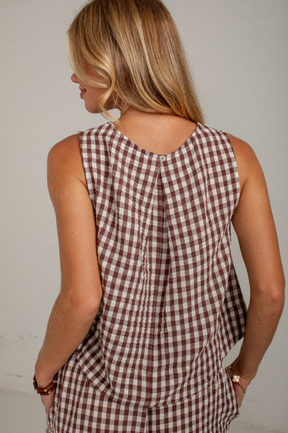 A woman shown from behind wearing a sleeveless brown and white gingham check top, photographed against a plain light wall.