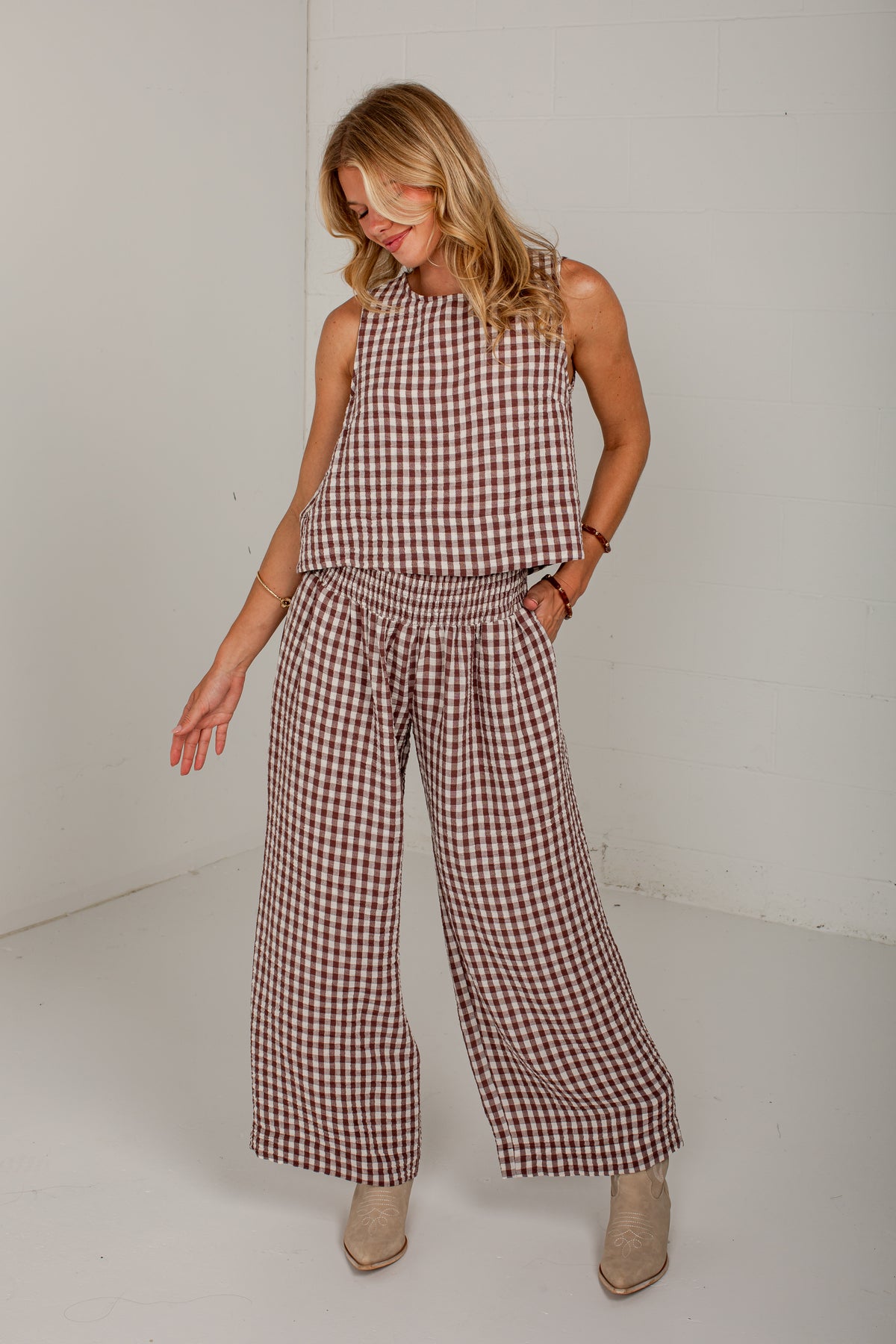 A woman modeling a sleeveless gingham jumpsuit in brown and white check, paired with beige ankle boots, standing against a white studio background.