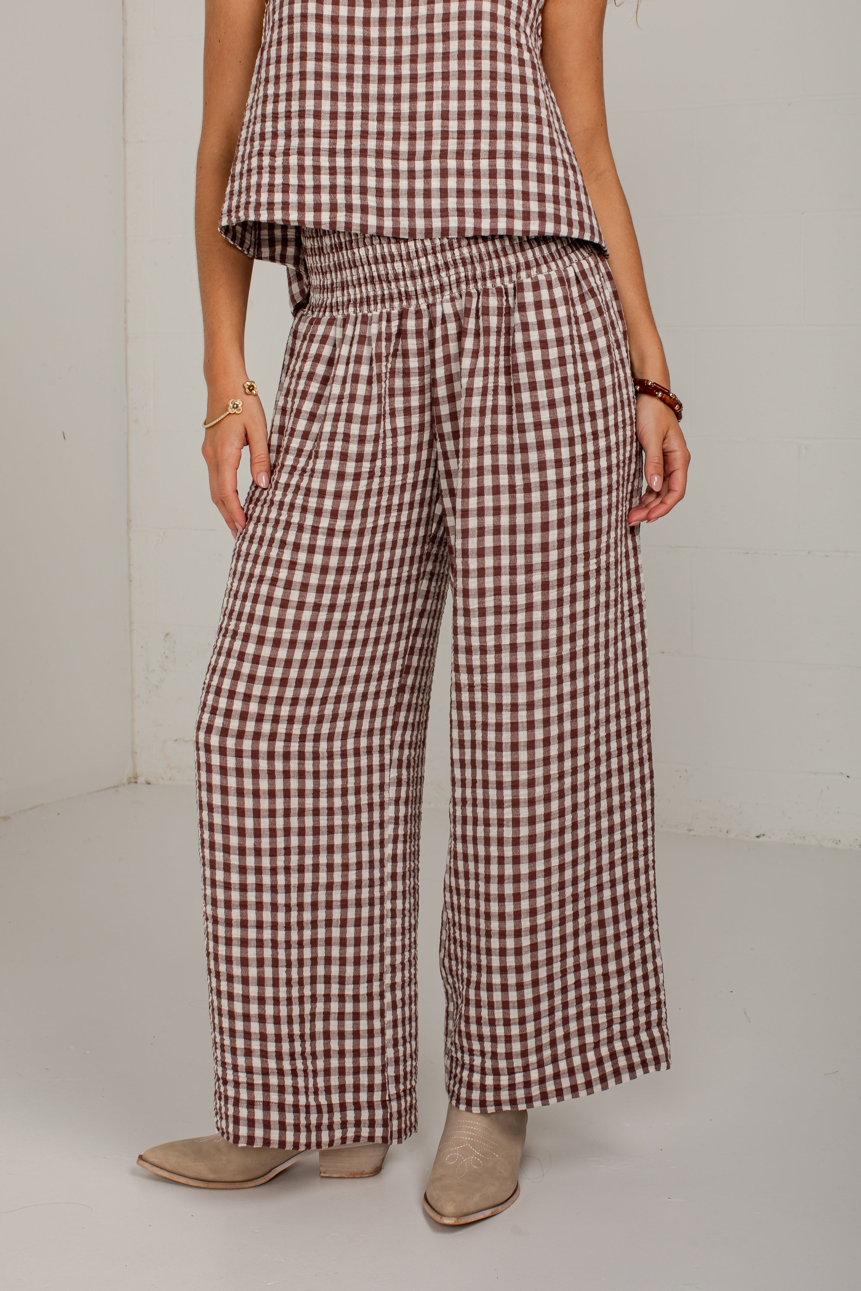 A model wearing brown and white gingham wide-leg pants with an elastic waistband, paired with a matching gingham top and beige boots, standing against a white wall.