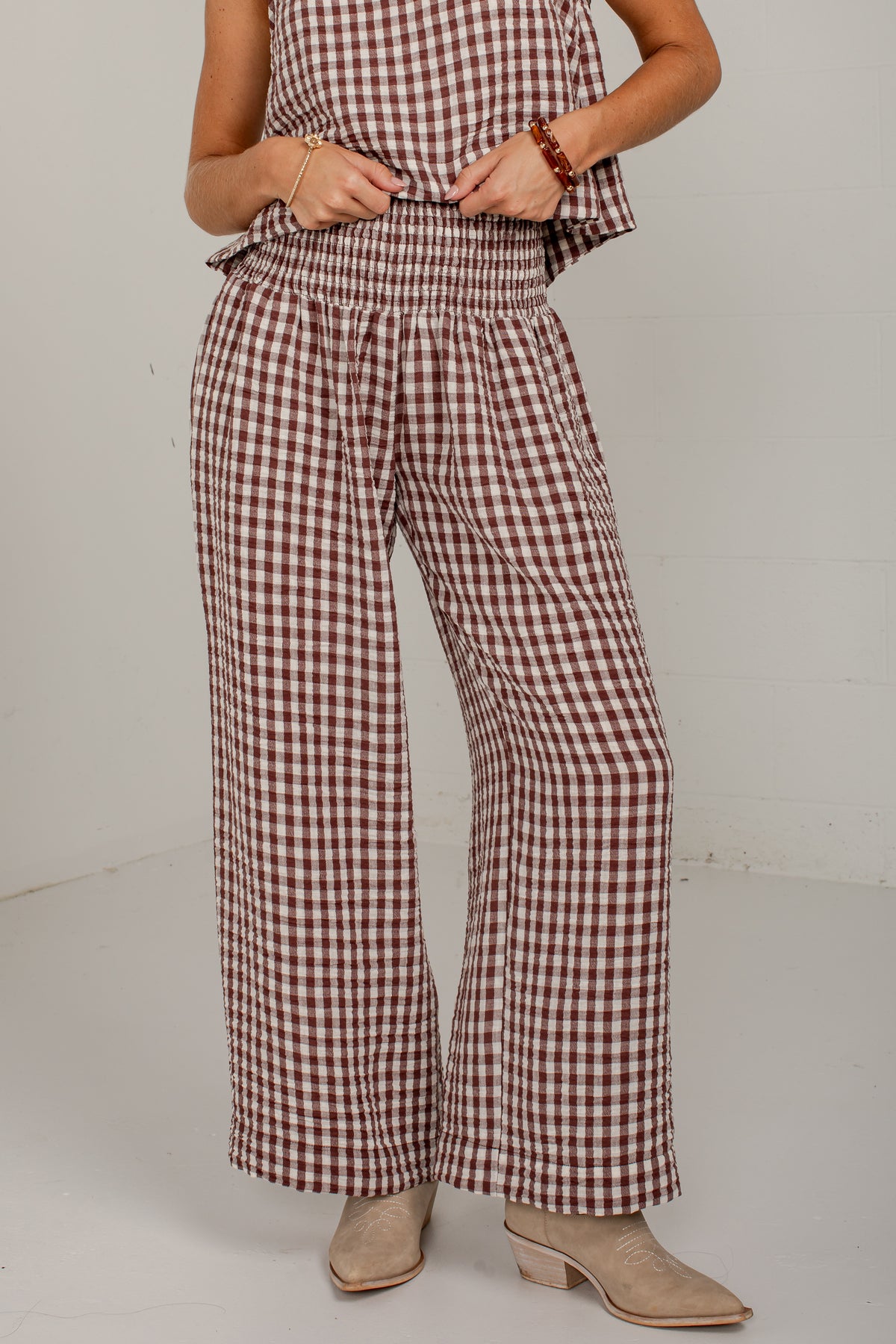 A person wearing brown and white gingham wide-leg pants with a smocked waistband, paired with beige ankle boots against a plain light gray studio background.