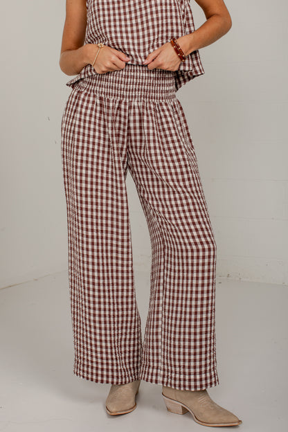 A person wearing brown and white gingham wide-leg pants with a smocked waistband, paired with beige ankle boots against a plain light gray studio background.