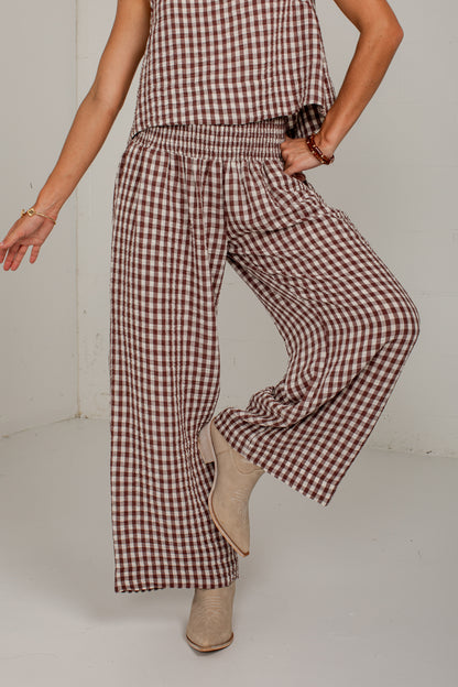 A model wearing a brown and white gingham check two-piece set consisting of a sleeveless top and wide-leg pants, paired with beige ankle boots.