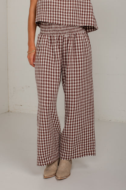 A person wearing brown and white gingham wide-leg pants with an elastic waistband, paired with beige ankle boots, photographed in a plain indoor setting.