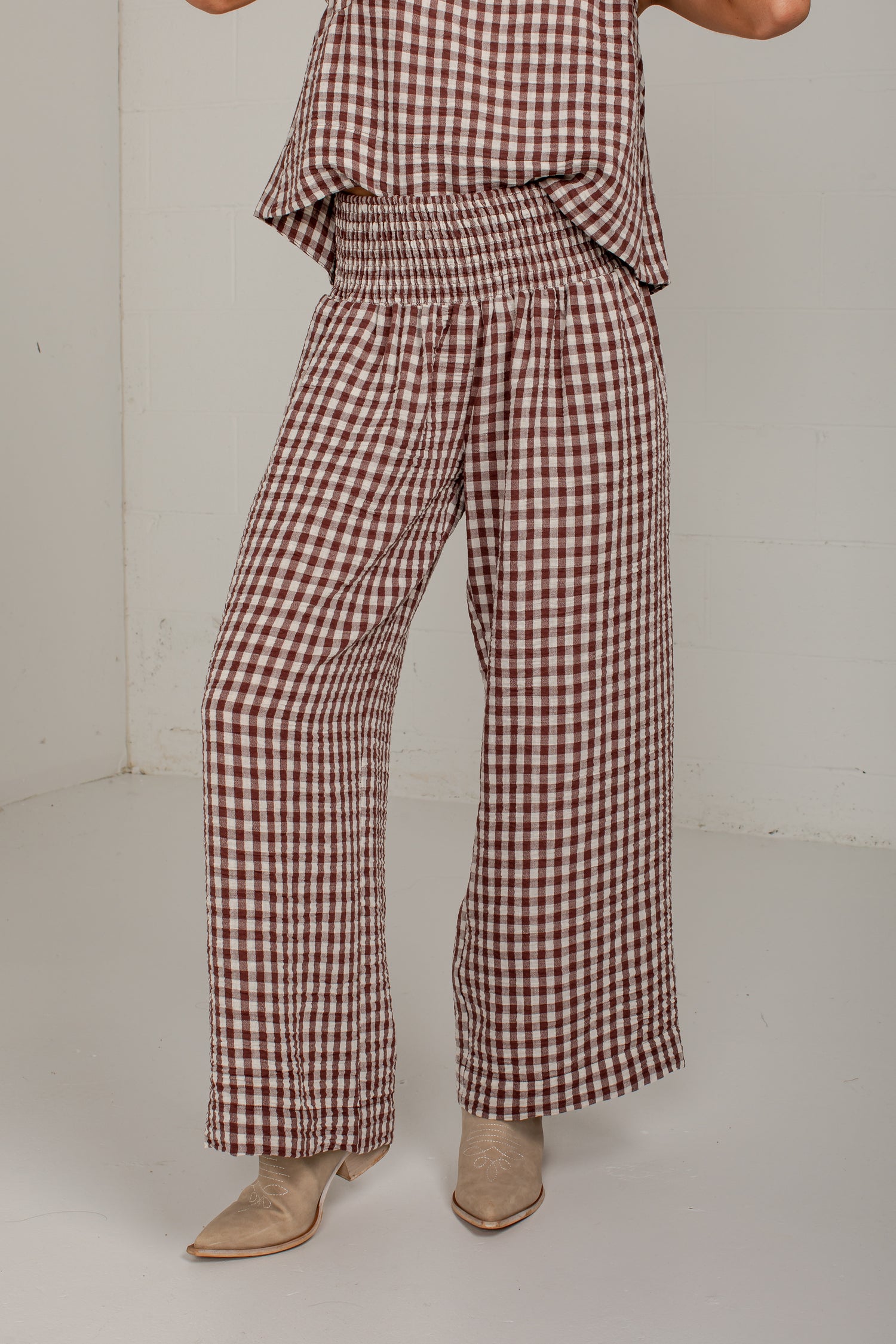 A person wearing brown and beige gingham wide-leg pants with a smocked elastic waist, paired with beige pointed-toe heeled boots, photographed in a plain studio corner.