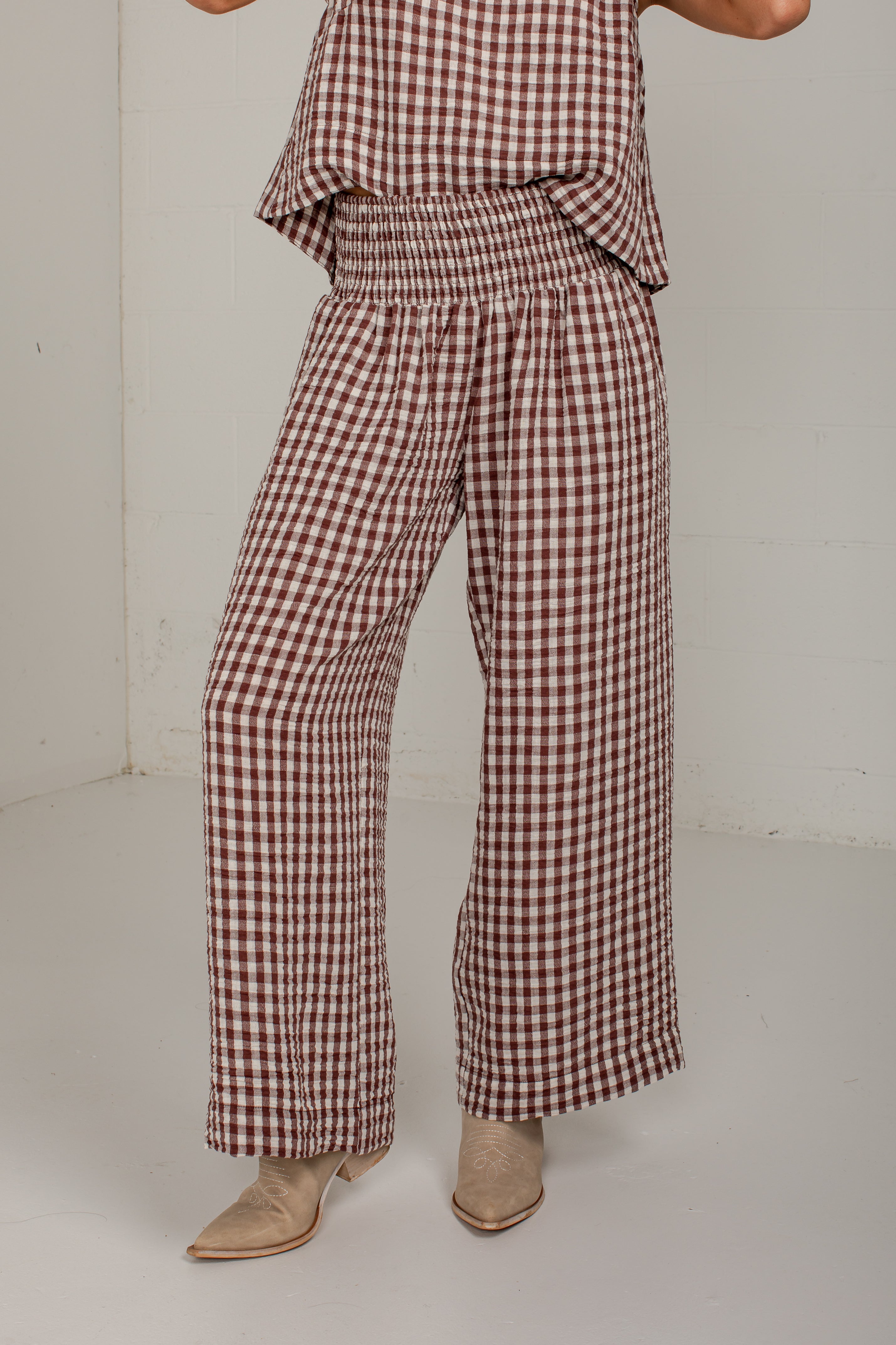 A person wearing brown and beige gingham wide-leg pants with a smocked elastic waist, paired with beige pointed-toe heeled boots, photographed in a plain studio corner.