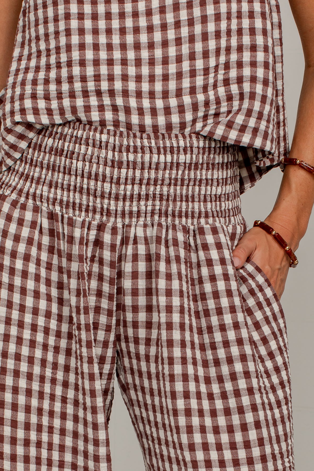Close-up of a person wearing a brown and white gingham check romper with an elastic waistband and side pockets, paired with a matching top.