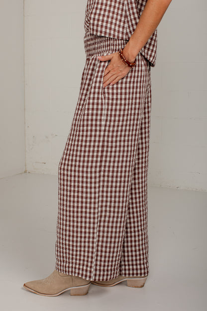 A person wearing brown and beige gingham wide-leg pants with a matching top, paired with beige pointed-toe boots, standing against a white wall.