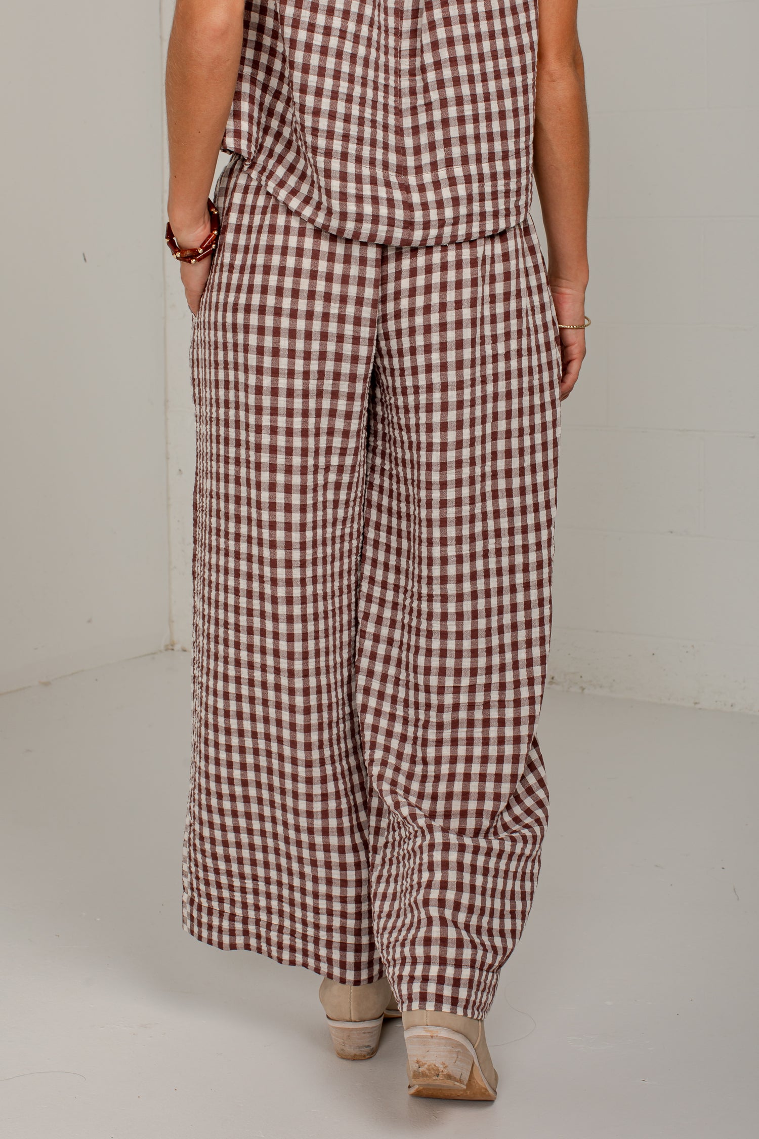 A person wearing a brown and white gingham check wide-leg jumpsuit, shown from the back in a studio setting.