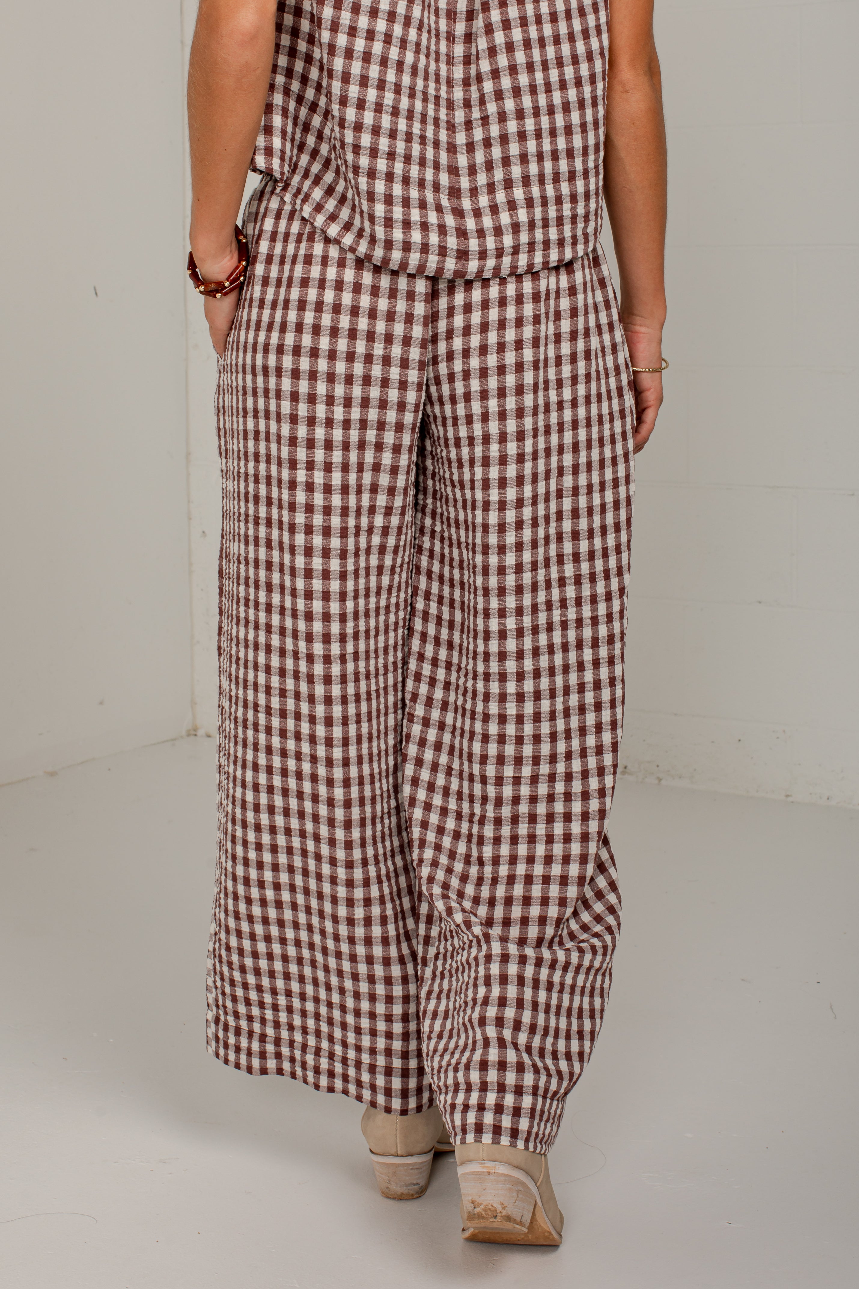 A person wearing a brown and white gingham check wide-leg jumpsuit, shown from the back in a studio setting.