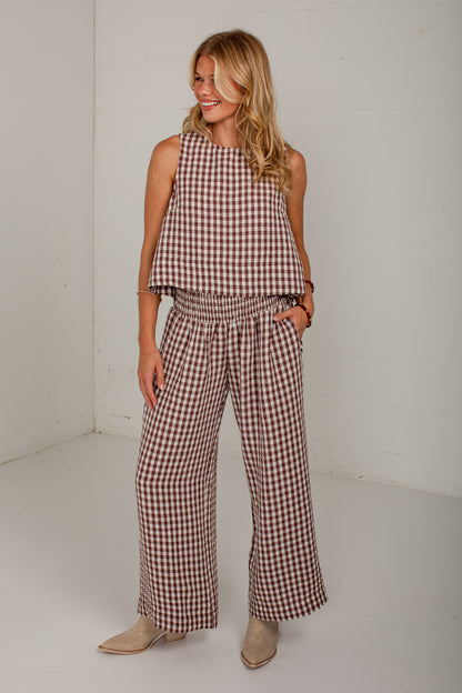 A woman modeling a sleeveless gingham top and matching wide-leg pants in a brown and white check pattern, standing against a plain white studio wall.