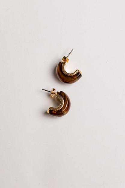 A pair of crescent-shaped hoop earrings with a glossy brown and amber marbled pattern and gold-tone metal borders, displayed on a clean off-white background.