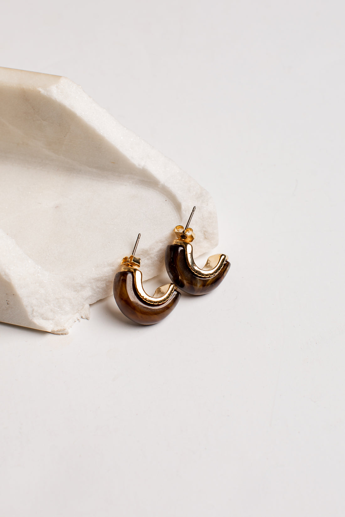 A pair of crescent-shaped hoop earrings with a glossy dark brown and gold finish, resting on a textured white stone prop against a clean white background.