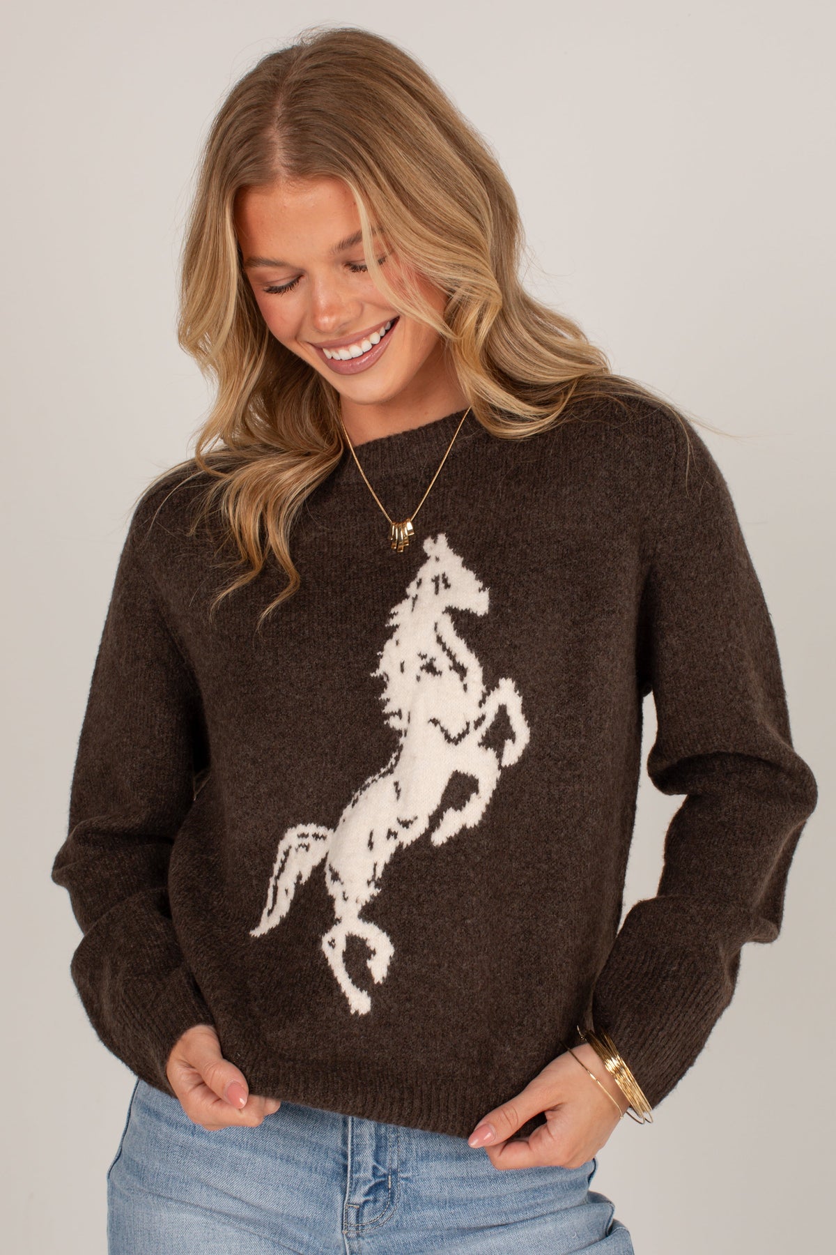 A woman wearing a dark brown knit sweater with a large white rearing horse graphic on the front, paired with light blue jeans, against a plain light gray background.