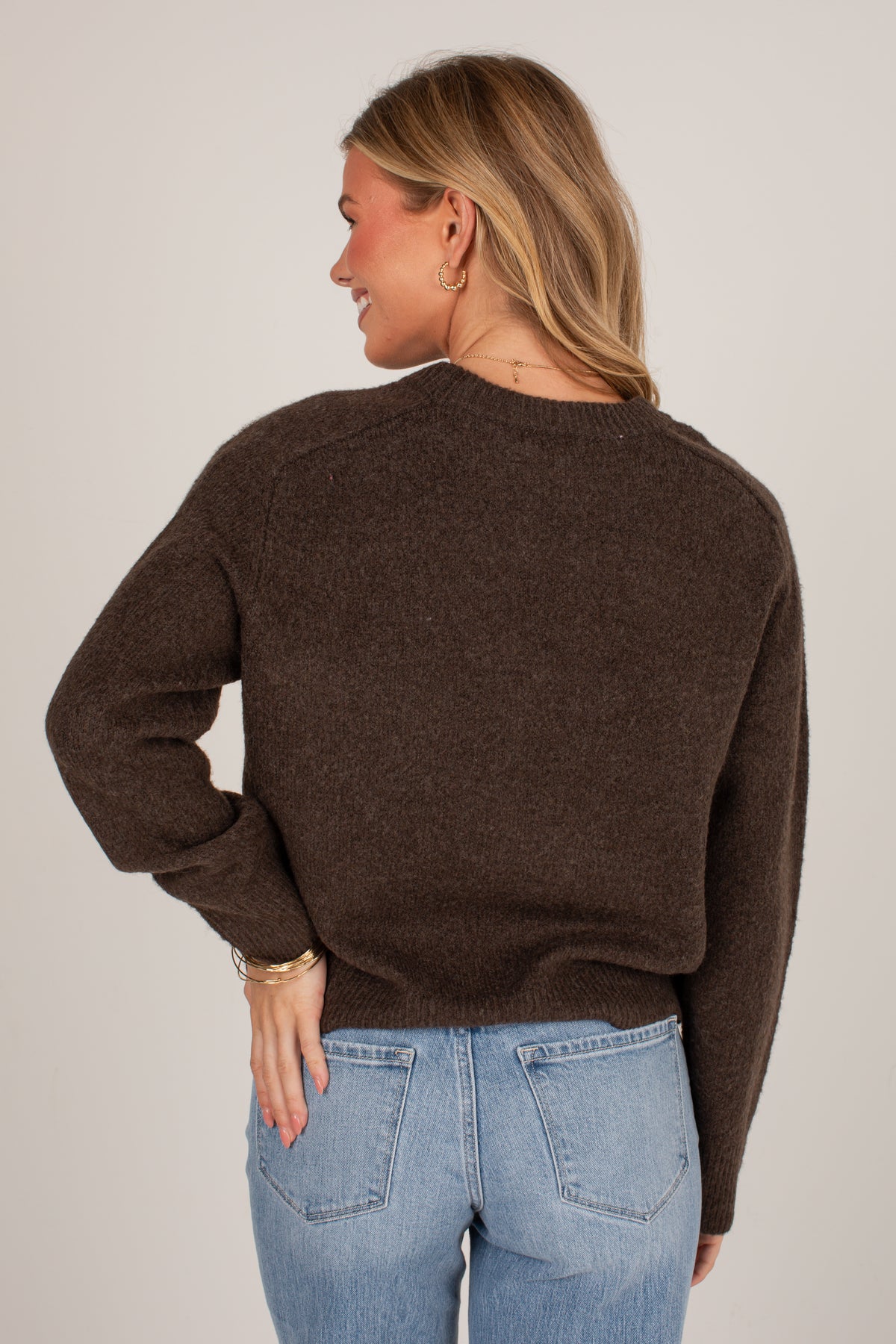 A woman shown from the back wearing a dark brown knit sweater and light blue jeans against a plain light gray studio background.