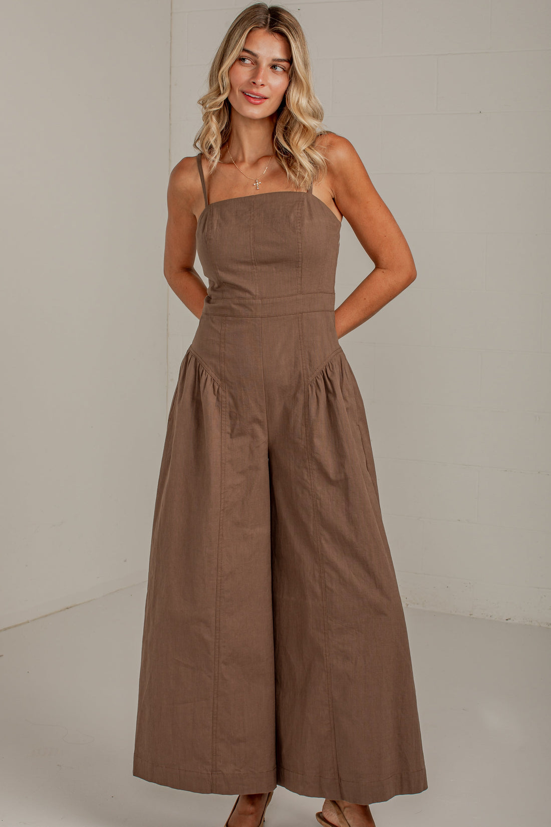 A woman modeling a brown wide-leg strapless jumpsuit in a bright, minimalist studio setting.
