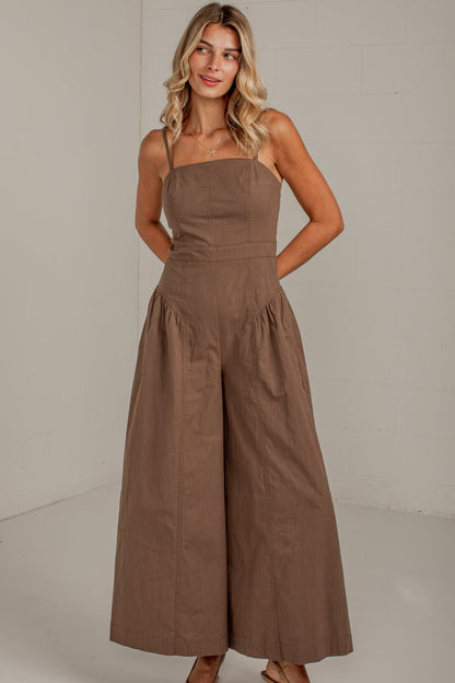 A woman modeling a brown wide-leg strapless jumpsuit in a bright, minimalist studio setting.