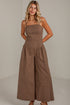 A woman modeling a brown wide-leg strapless jumpsuit in a bright, minimalist studio setting.
