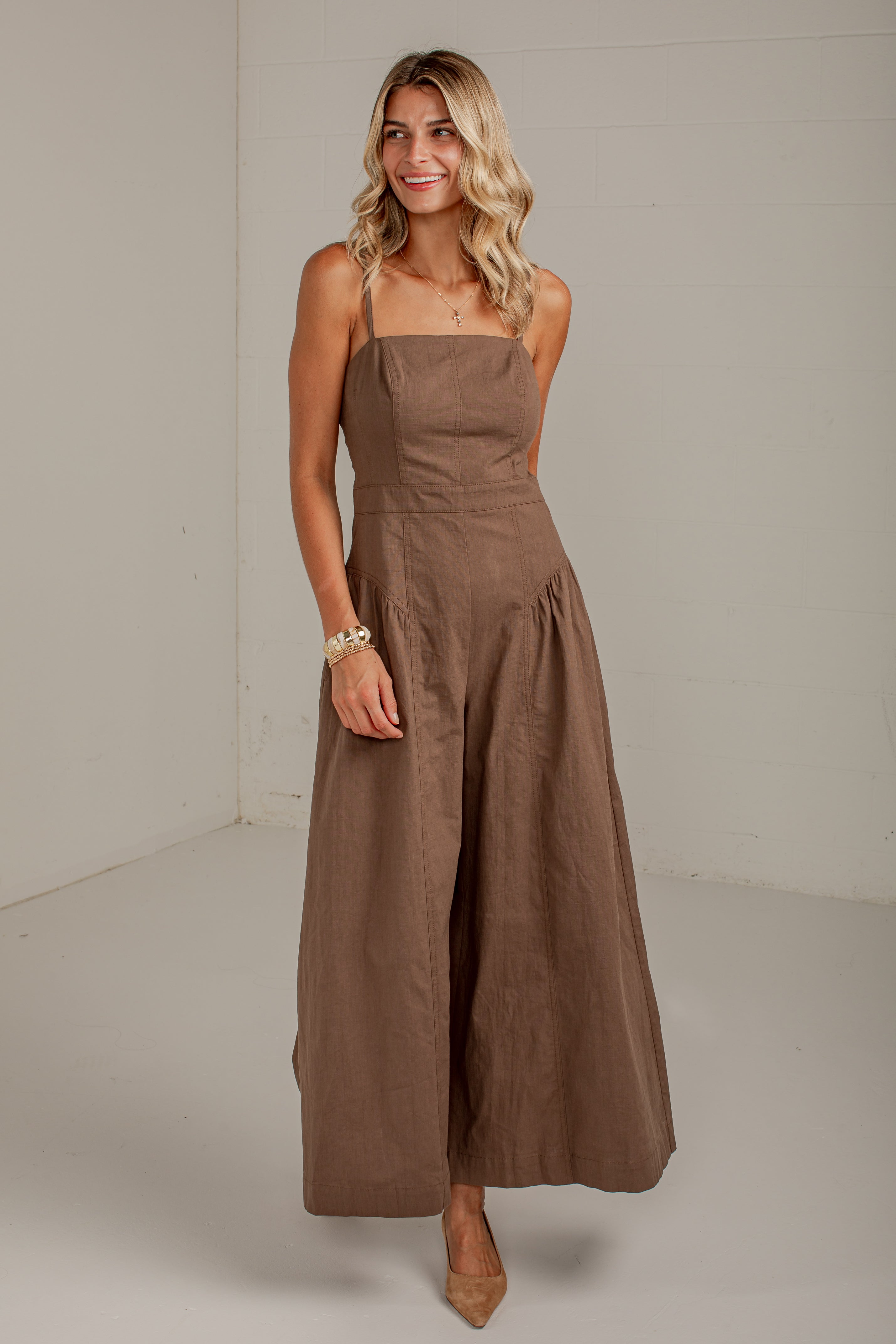 A woman modeling a brown wide-leg jumpsuit with thin straps and a square neckline, standing in a neutral studio setting.