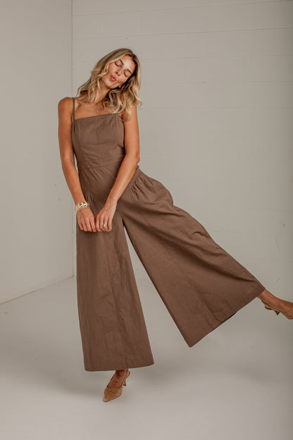 A woman modeling a brown wide-leg jumpsuit with a strapless bodice, standing in a studio with a light gray background.