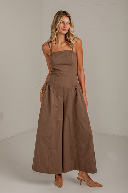A woman modeling a brown wide-leg jumpsuit with thin straps and a gathered waist, standing against a plain light gray studio wall.