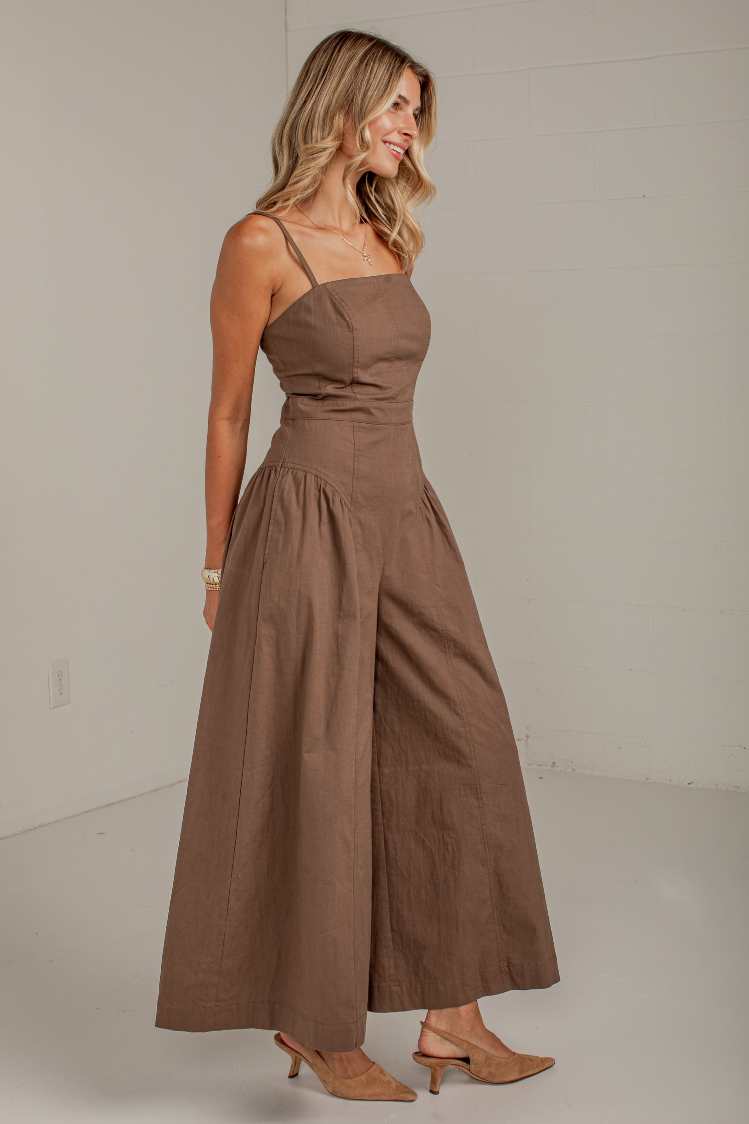 A woman modeling a brown sleeveless maxi dress with thin straps, a fitted bodice, and wide legs, standing in a plain indoor studio setting.