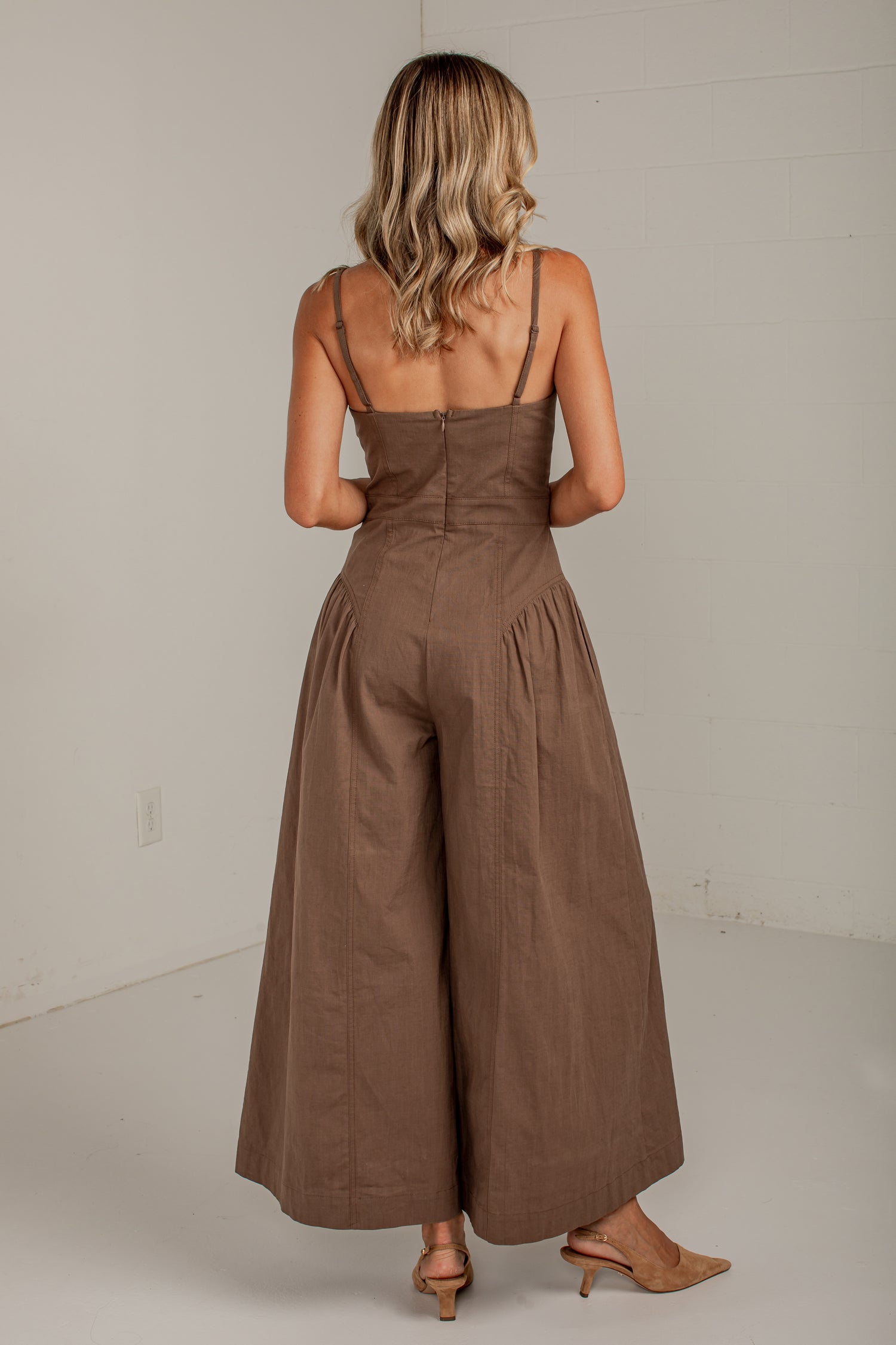 A woman viewed from behind wearing a brown sleeveless jumpsuit with thin shoulder straps, a fitted waist, and wide legs, standing in a plain indoor room with light neutral walls.