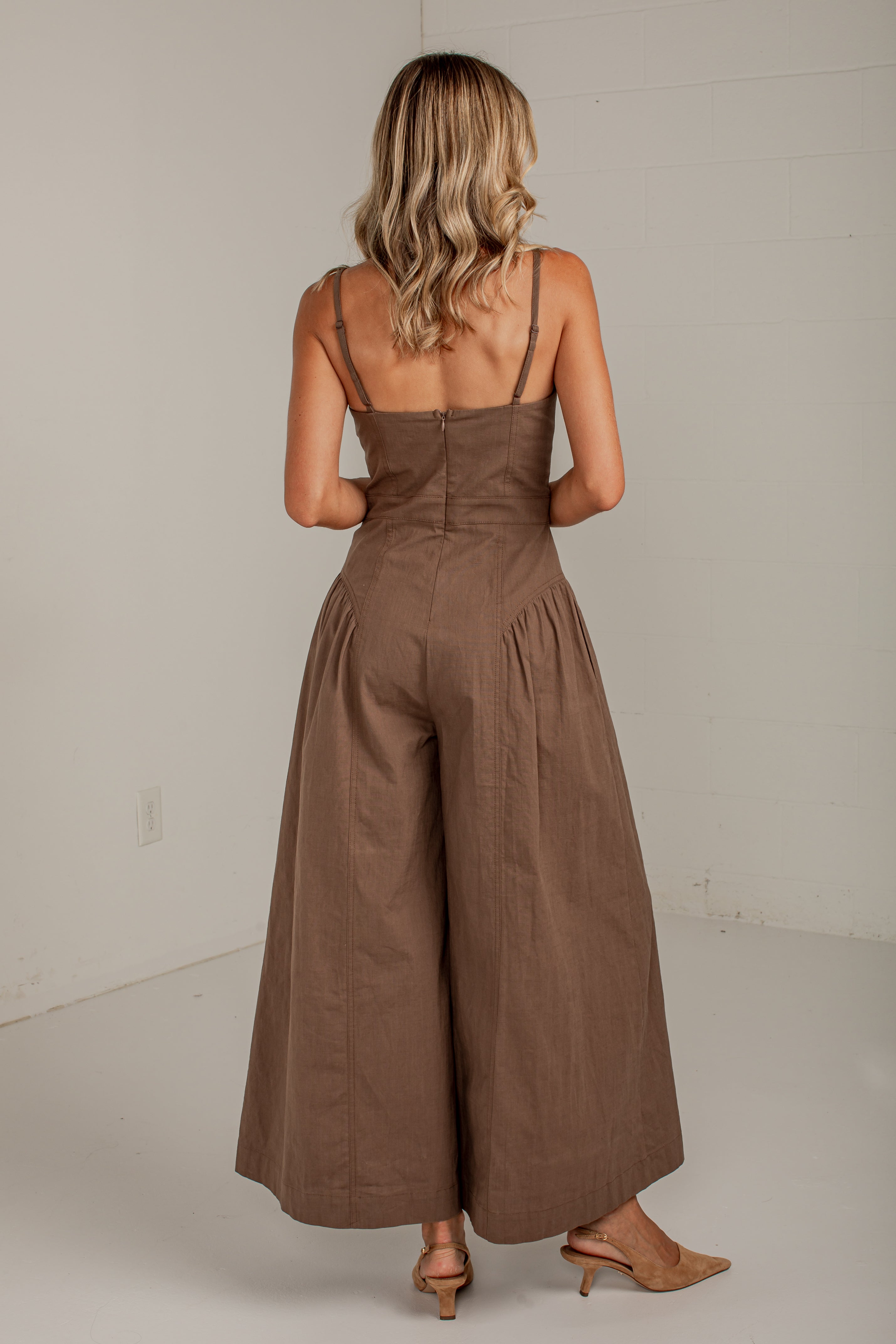 A woman viewed from behind wearing a brown sleeveless jumpsuit with thin shoulder straps, a fitted waist, and wide legs, standing in a plain indoor room with light neutral walls.