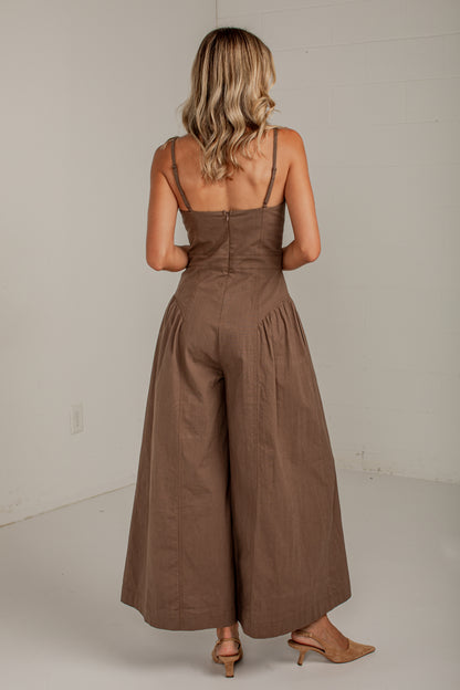 A woman viewed from behind wearing a brown sleeveless jumpsuit with thin shoulder straps, a fitted waist, and wide legs, standing in a plain indoor room with light neutral walls.